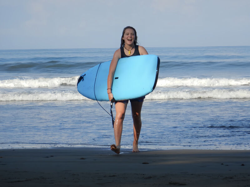 A young woman is walking on a beach, carrying a light blue surfboard under her arm. She is smiling and appears to be enjoying herself. The ocean is behind her with small waves, and the sky is bright. The sand is visible in the foreground, and the overall scene suggests a sunny day at the beach.